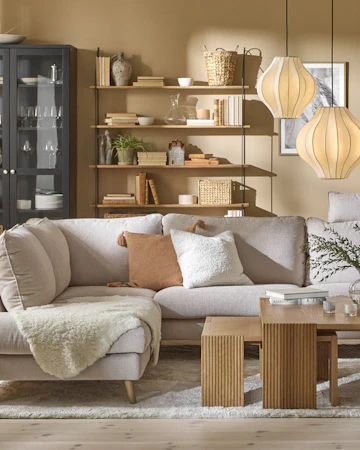 I ett gult vardagsrum står en ljusgrå soffa framför ett svart vitrinskåp och en vägghylla med hyllplan i trä