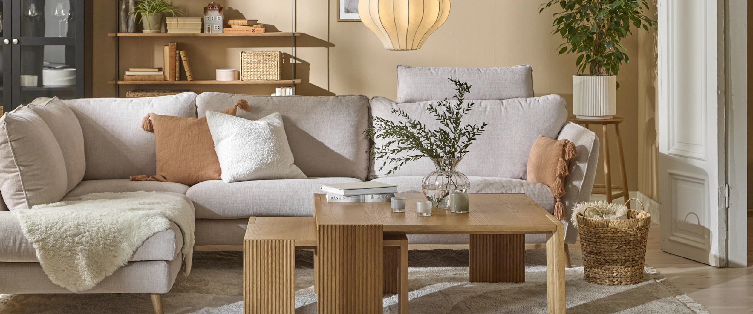 I ett gult vardagsrum står en ljusgrå soffa framför ett svart vitrinskåp och en vägghylla med hyllplan i trä