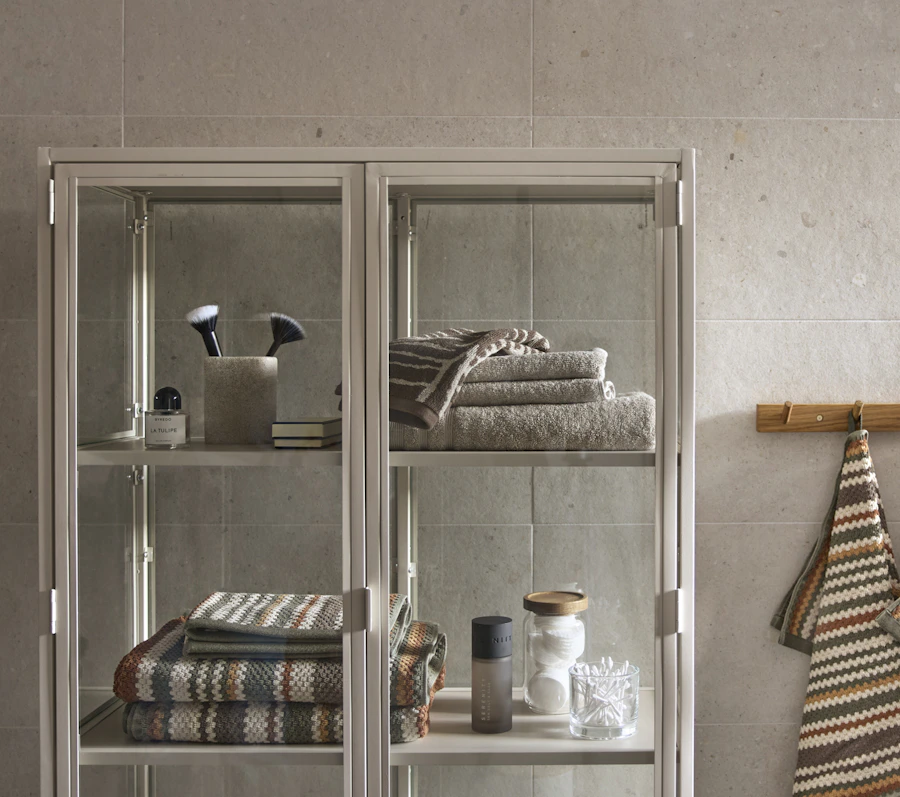 I ett badrum står ett skåp i metall och glas fylld med handdukar och andra badrumsatteraljer