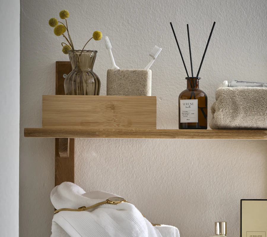 På en hylla i ek står en diffuser och en förvaringsbox i trä med tandborstmugg och vas inuti