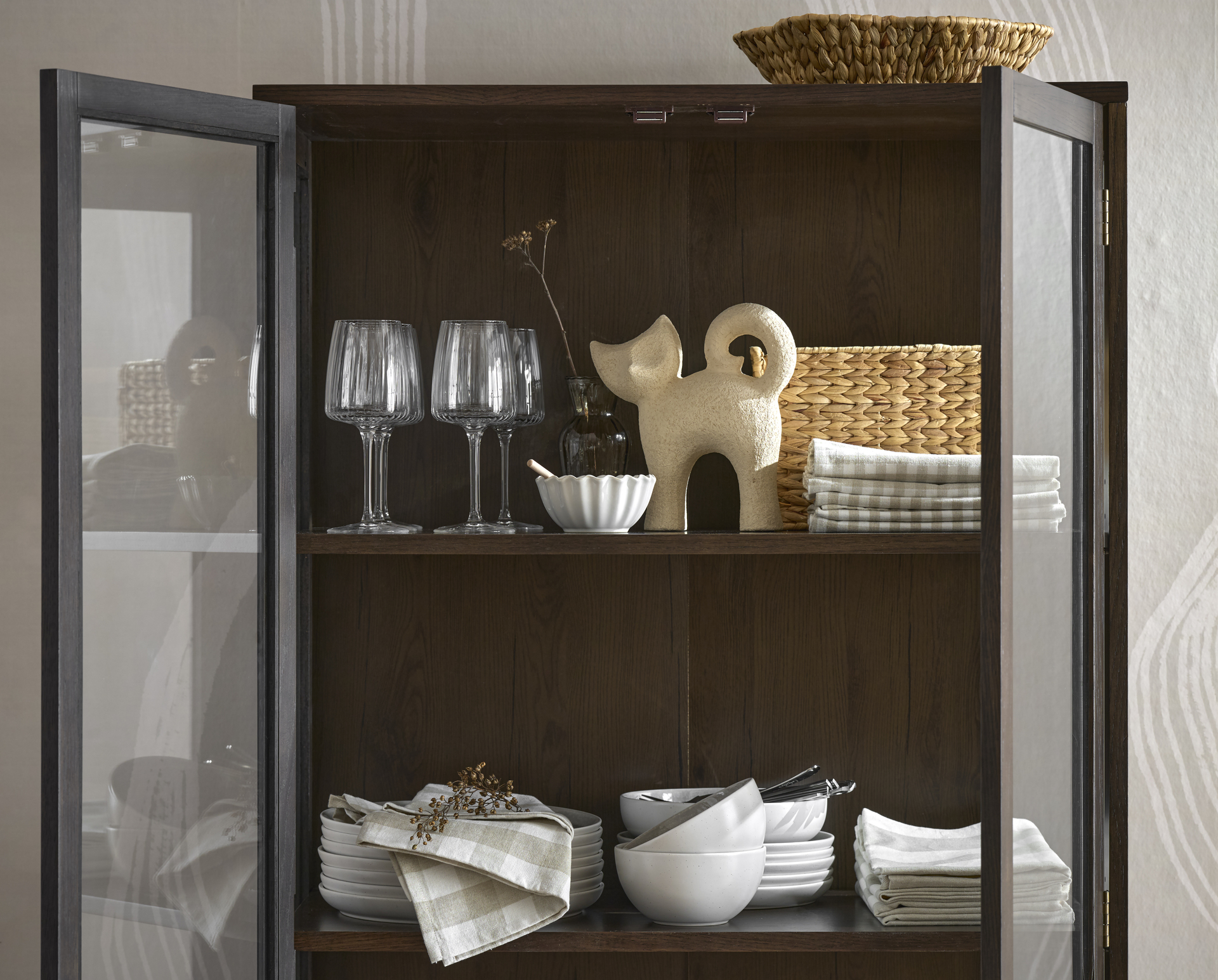 I ett öppet mörkbrunt vitrinskåp står en beige prydnadskatt, några vinglas och en vit servis.