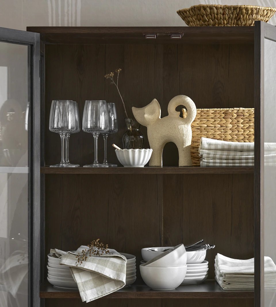 I ett öppet mörkbrunt vitrinskåp står en beige prydnadskatt, några vinglas och en vit servis.