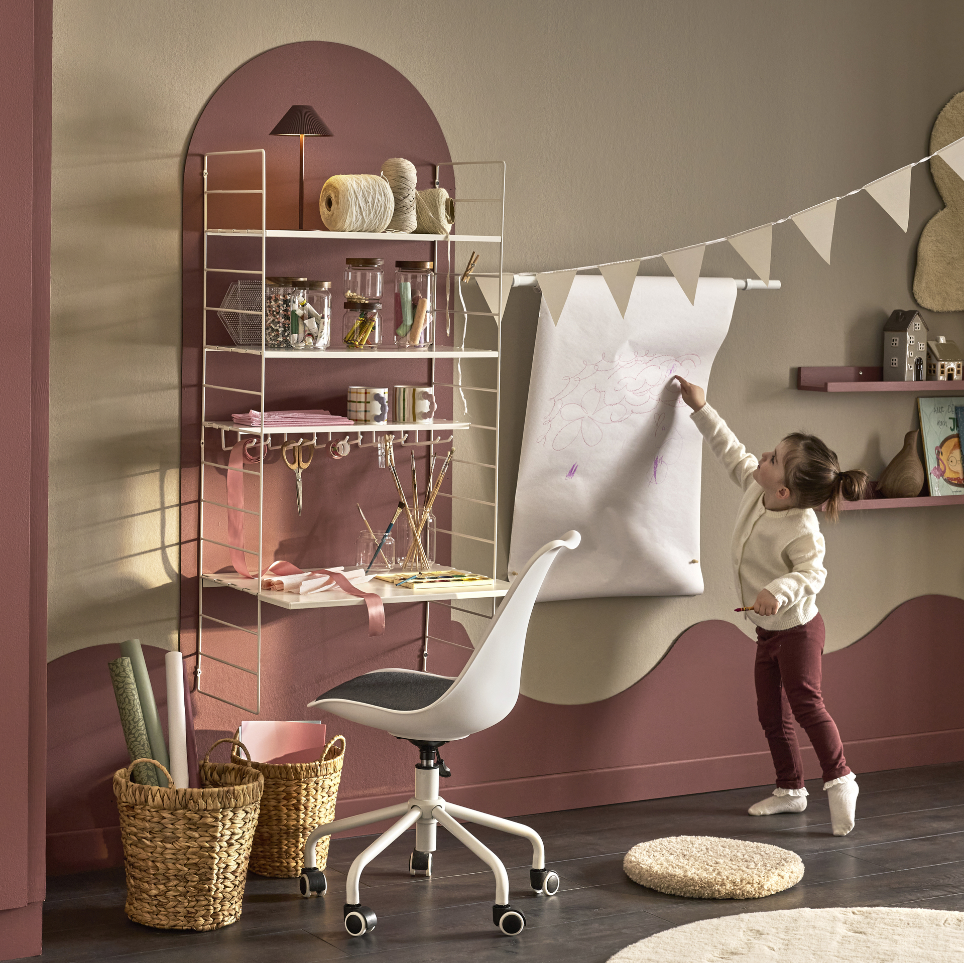 I ett barnrum målat i gult och rosa står en flicka bredvid ett skrivbord med en hylla över och målar
