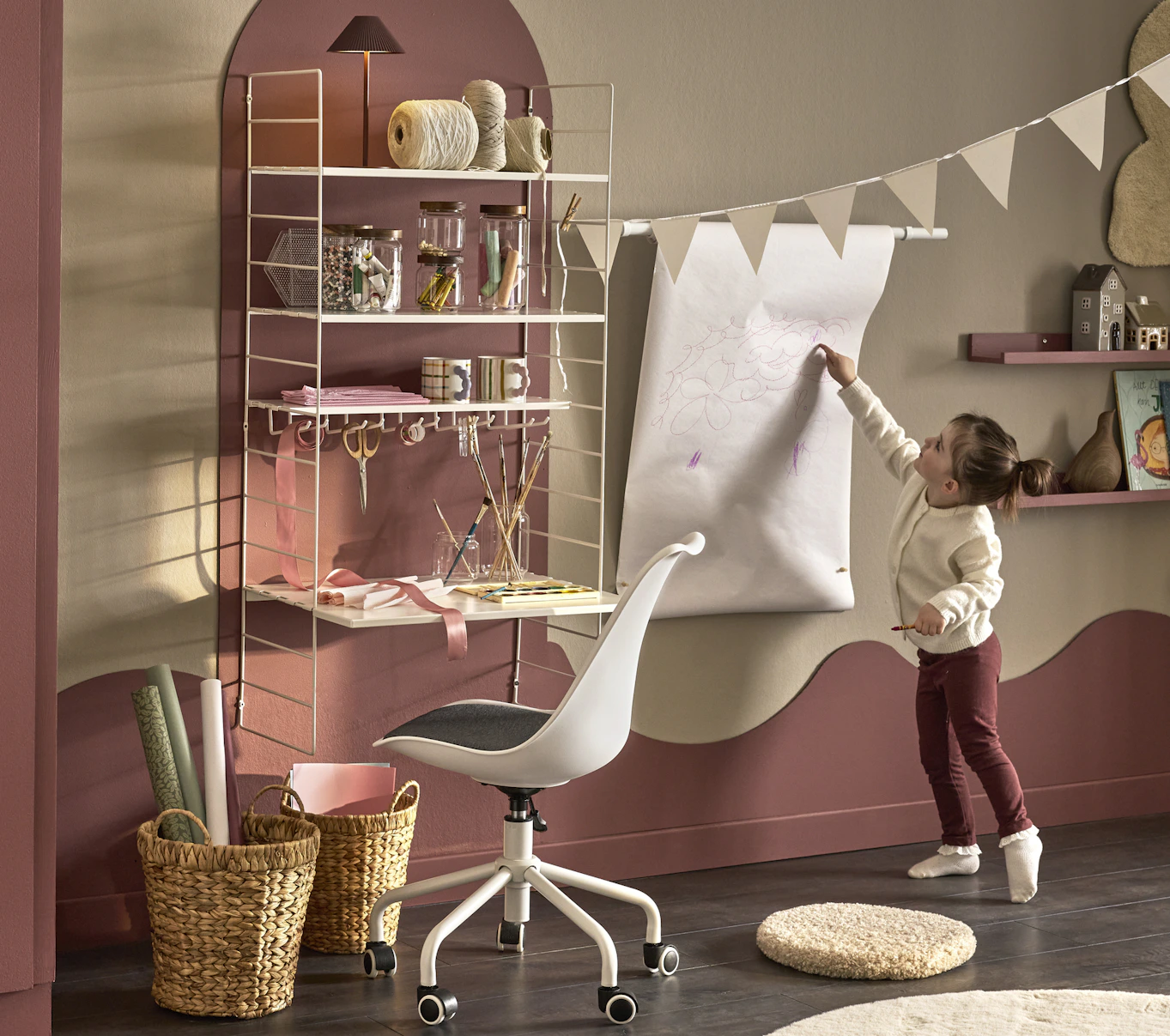 I ett barnrum målat i gult och rosa står en flicka bredvid ett skrivbord med en hylla över och målar