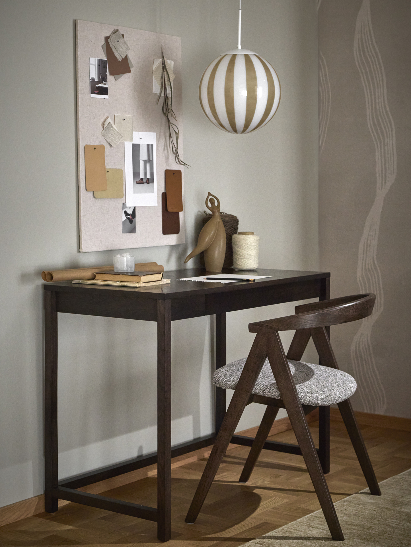 Ett mörkbrunt skrivbord och matchande stol står intill en grå vägg. Över skrivbordet hänger en rund taklampa med randigt mönster.