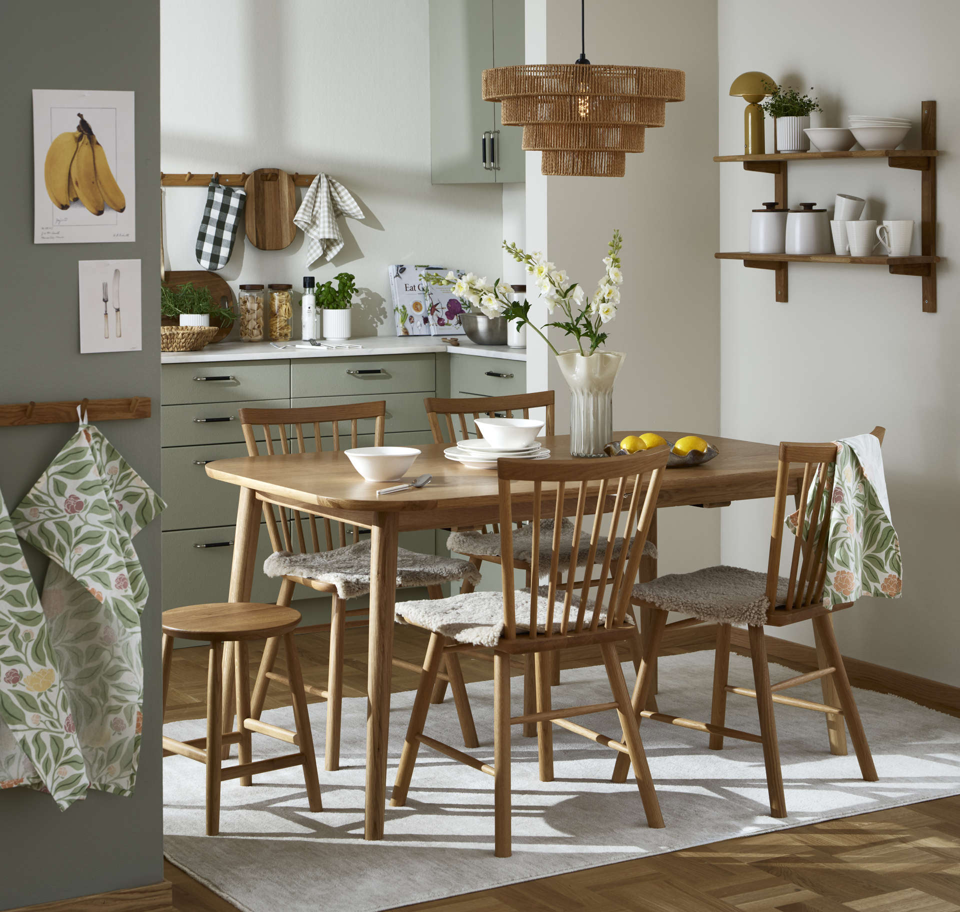 Mitt i ett beige kök står ett matbord och stolar i ek. I köket sittet en vägghylla med porslin och förvaringsburkar