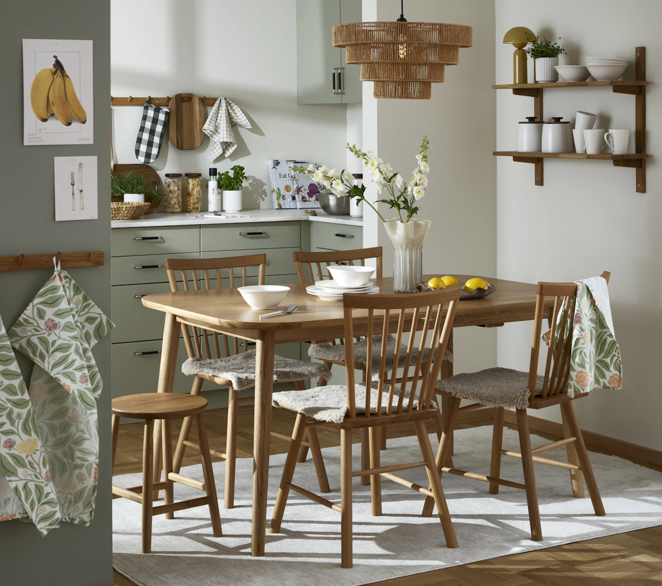 Mitt i ett beige kök står ett matbord och stolar i ek. I köket sittet en vägghylla med porslin och förvaringsburkar