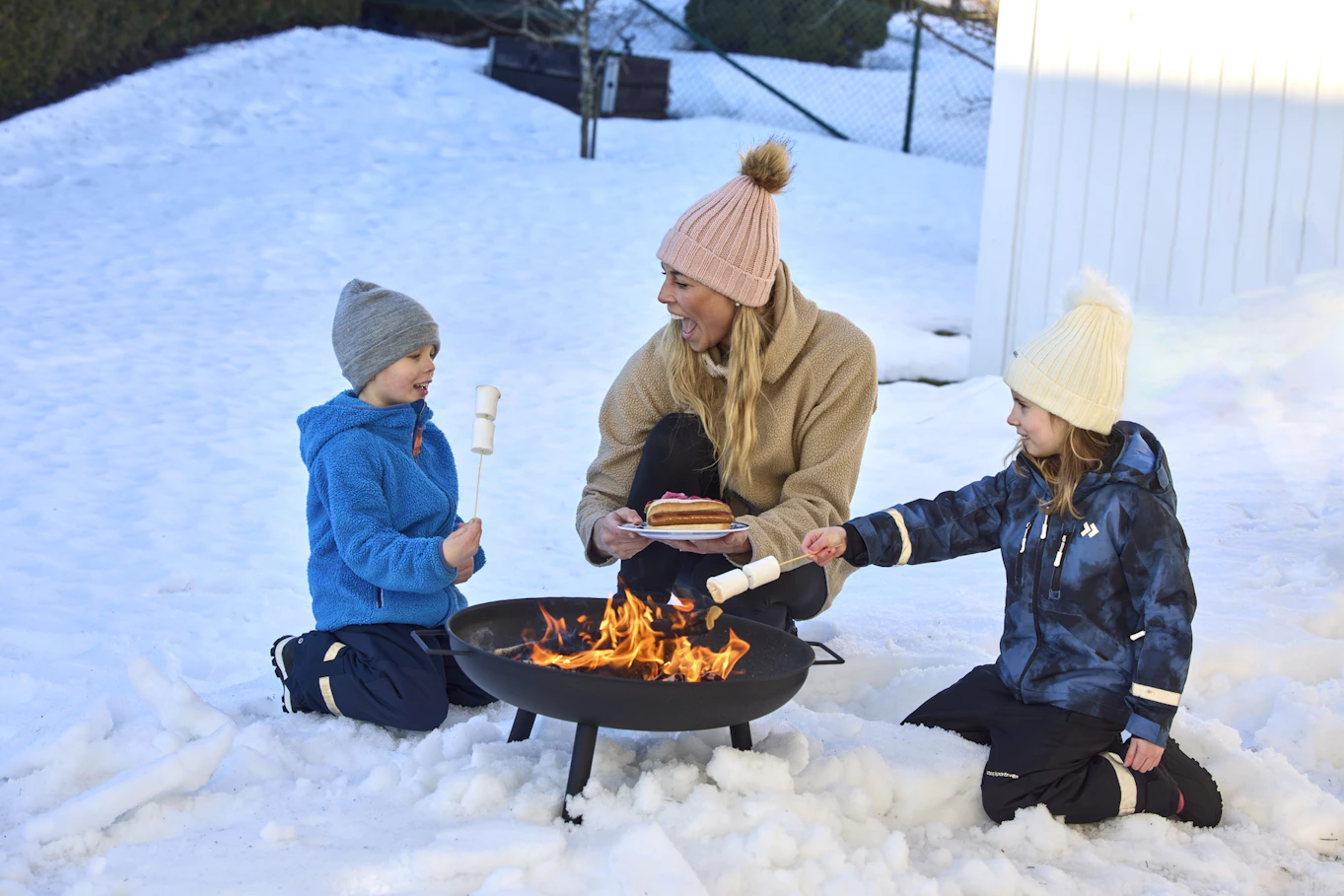 Kvinna sitter på huk i snö tillsammans med två små barn och grillar marshmallows över ett eldfat. 