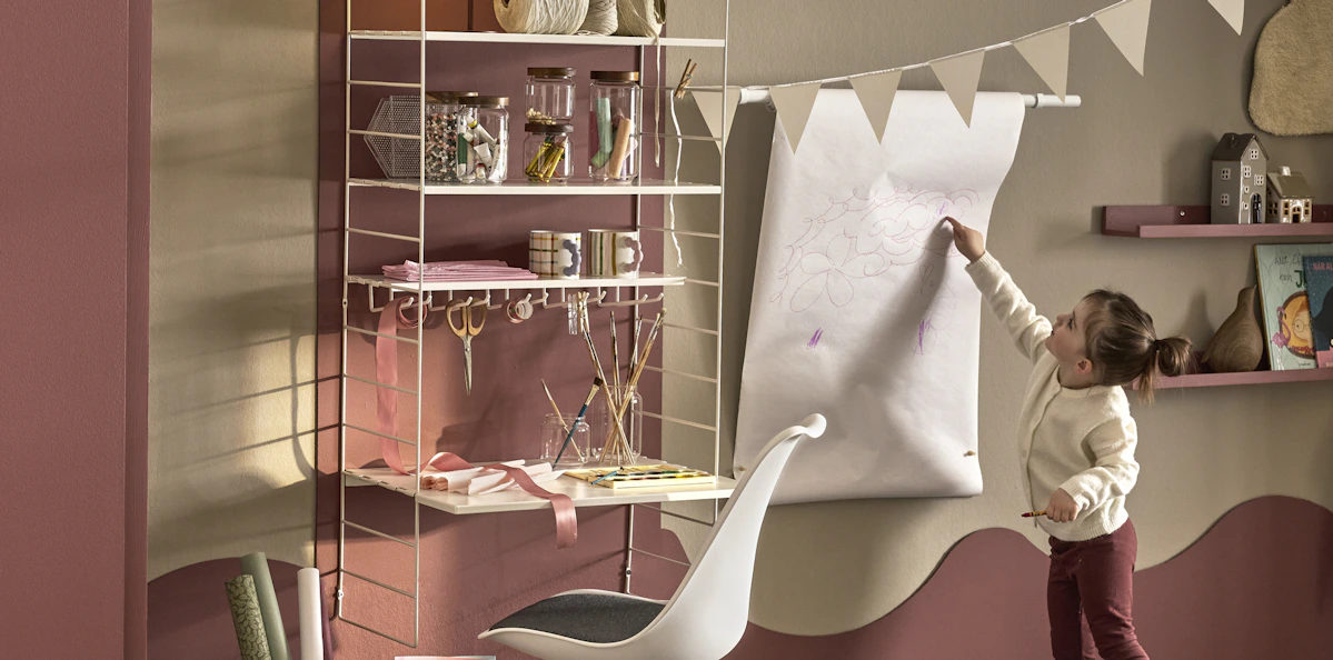 I ett rum står en flicka brevis ett skrivbord och målar