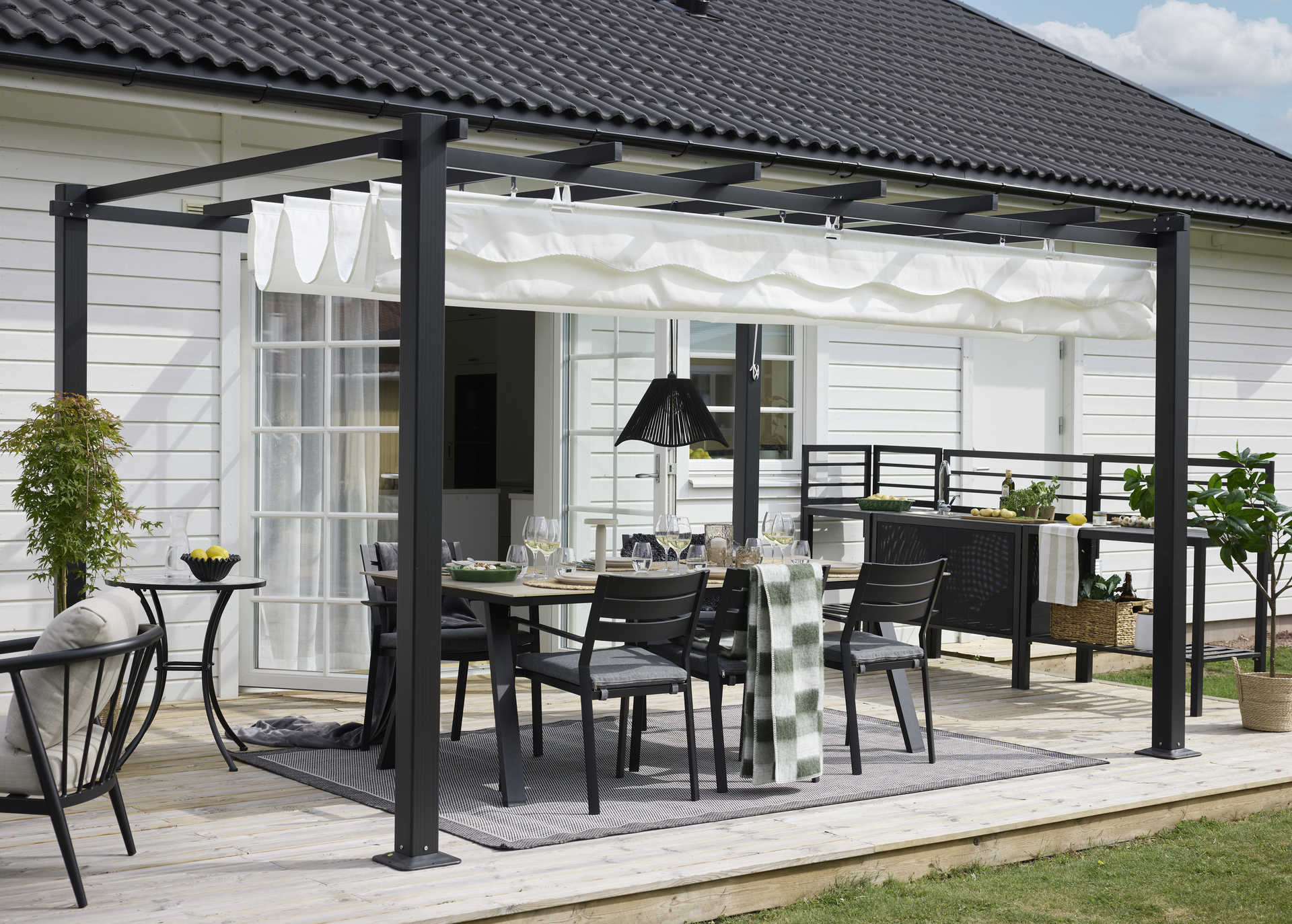 Utematgrupp under pergola i svart trä på terrass vid modern vila med svart tak och vita väggar.