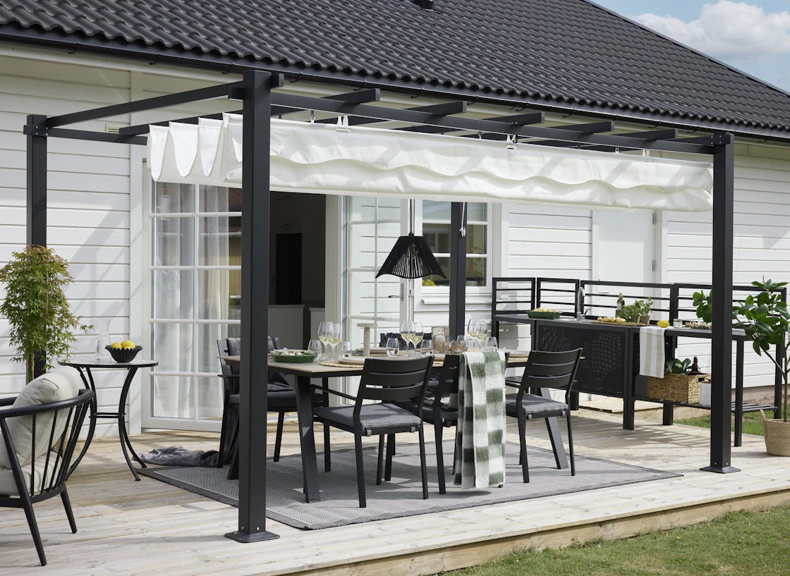 Utematgrupp under pergola i svart trä på terrass vid modern vila med svart tak och vita väggar.