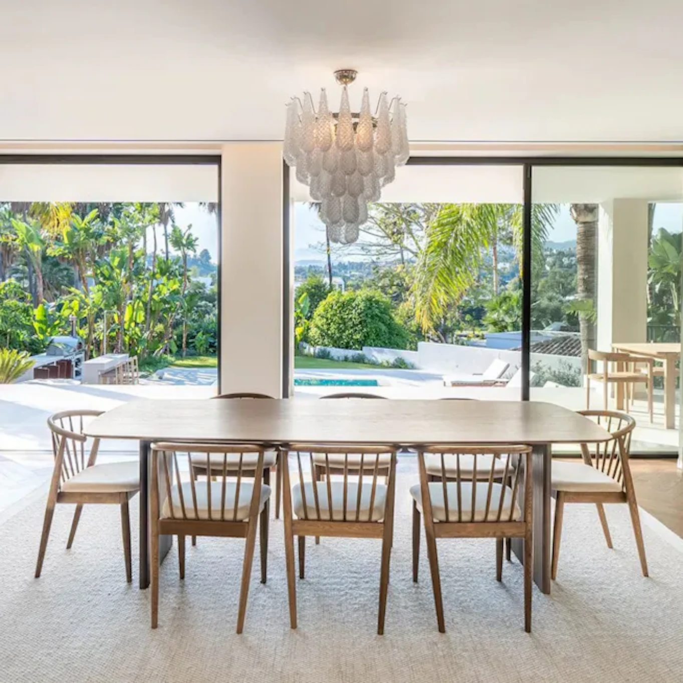 Matbord med ljus skiva och pinnstolar runtom står i ett luftigt och ljust vardagsrum med stora fönster som vetter ut mot medelhavsmiljö med pool och palmväxter. 
