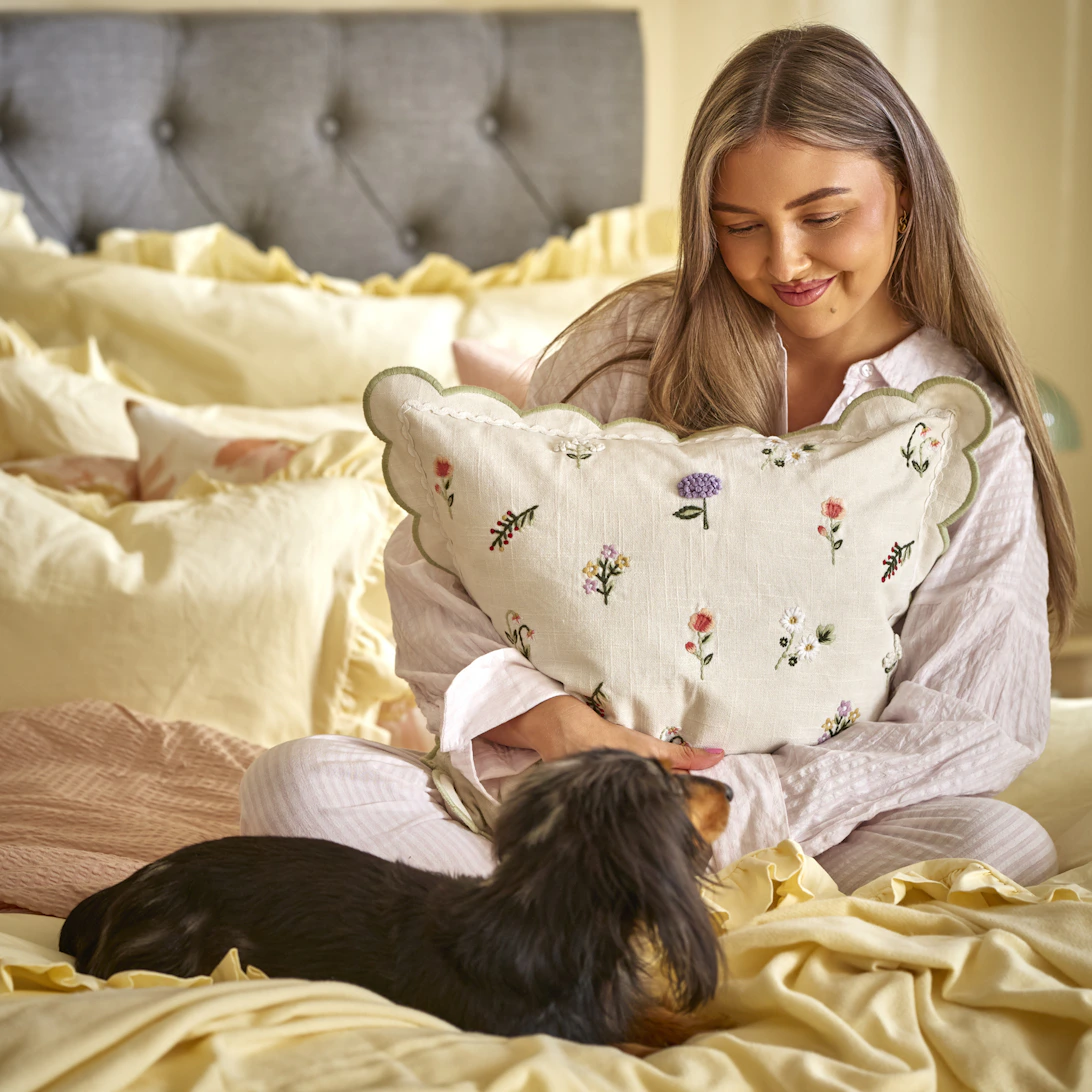 En tjej sitter i en säng bredvid en brun liten hund och kramar en kudde med blommönster på