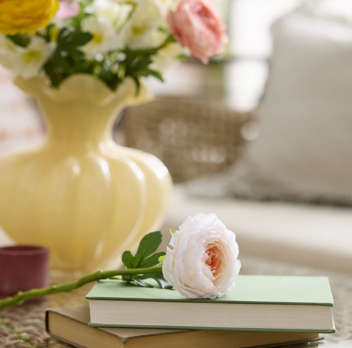 en ljusrosa konstväxt ligger på en bok på ett soffbord. I bakgrunden skymtas en gul vas