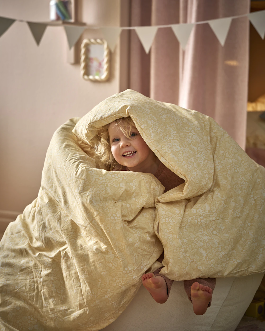 en blond flicka sitter i en säng invirad i ett täcke med gult påslakan