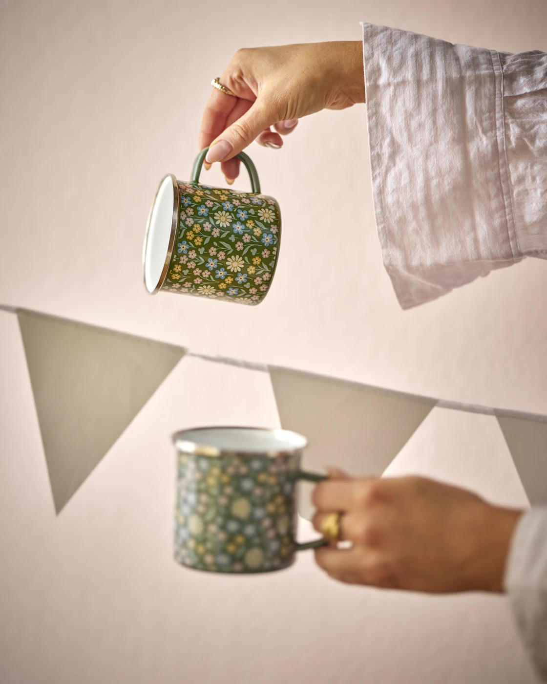 två händer håller fram två gröna blommiga muggar framför en rosa vägg
