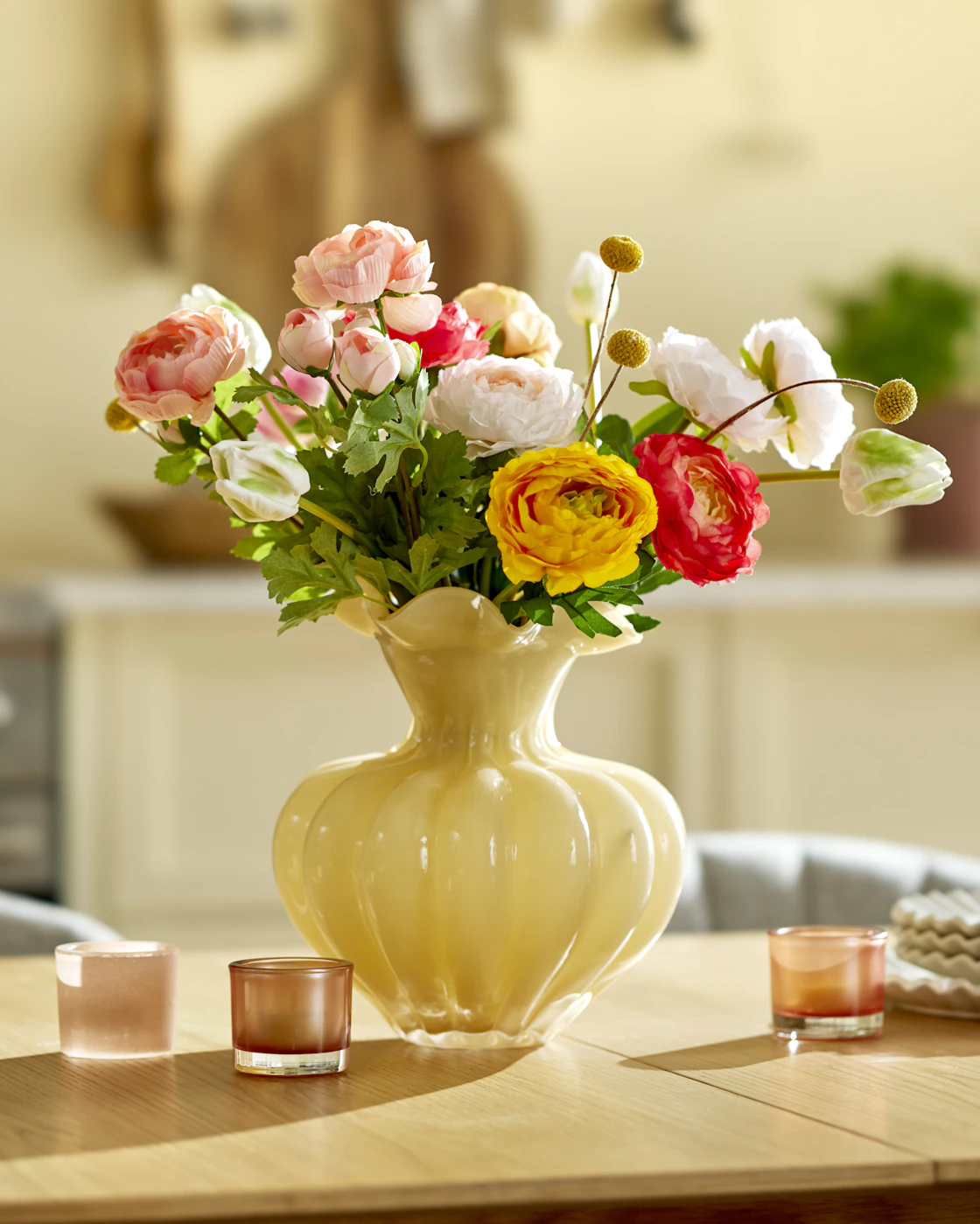 en gul vas med konstväxter i olika färger står på ett matbord i trä