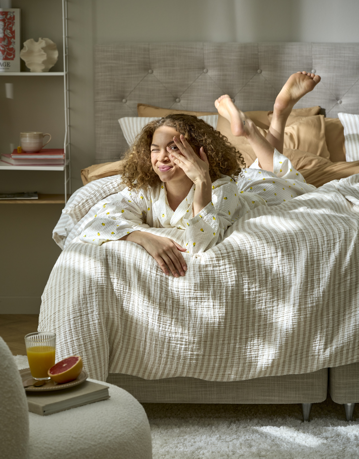 Ung kvinna med lockigt hår ligger i fotändan på säng och tittar in i kameran medan solljus tittar in genom skira gardiner. 