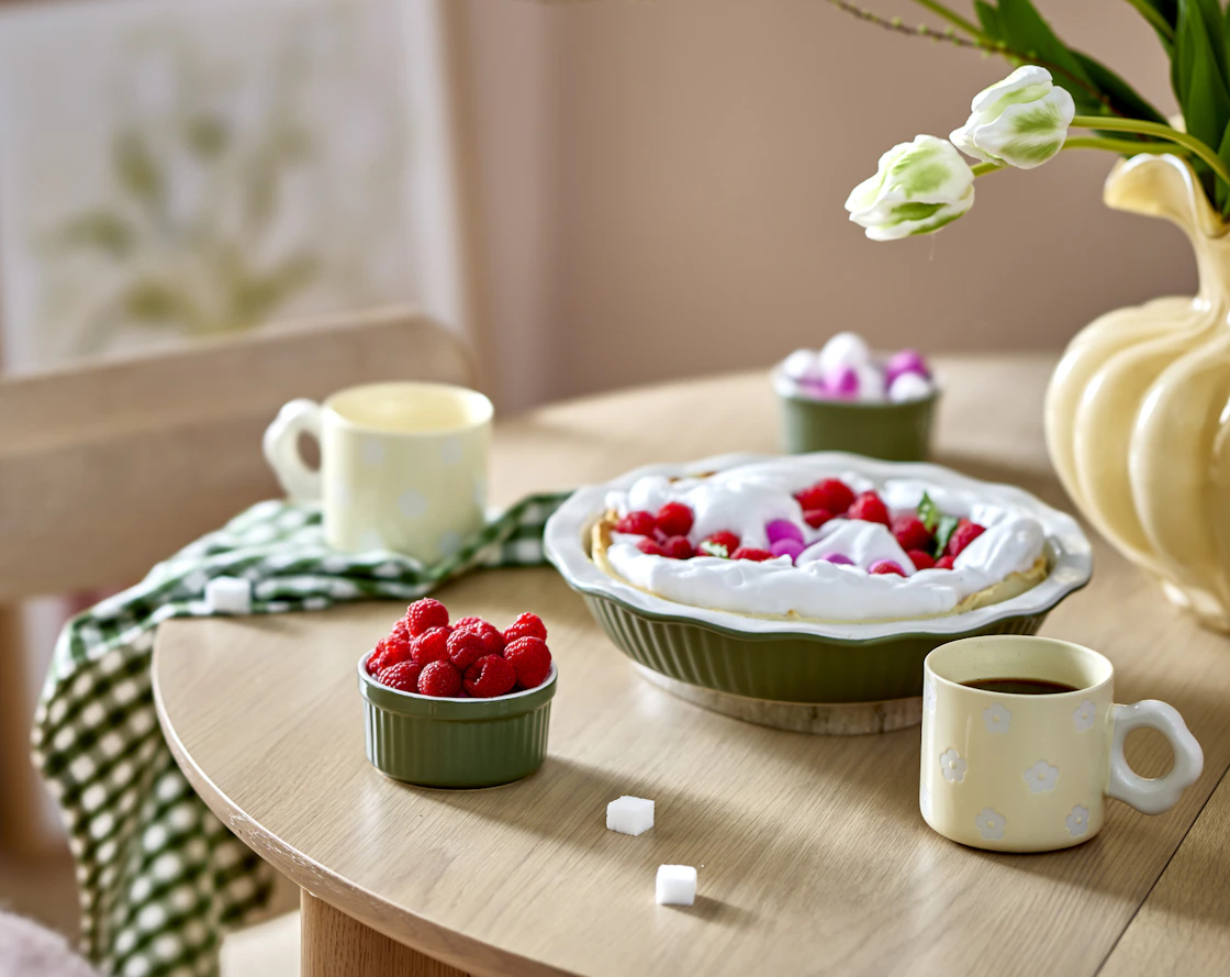 på ett bord i trä står en grön pajform, två gula muggar med blommor på och en liten grön skål med hallon