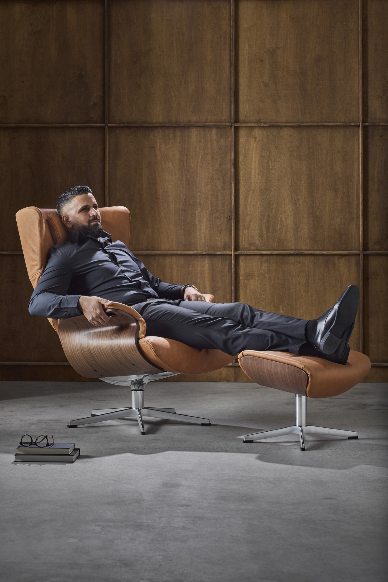I en brun skinnsfåtölj med underrede i valnöt sitter en man bakåtlutad med fötterna på en fotpall