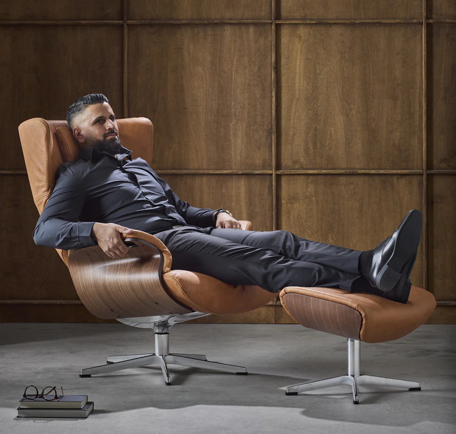 I en brun skinnsfåtölj med underrede i valnöt sitter en man bakåtlutad med fötterna på en fotpall