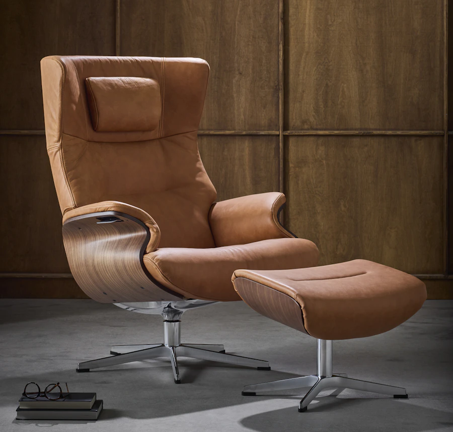 En brun skinnsfåtölj med underrede i valnöt står bredvid en fotpall