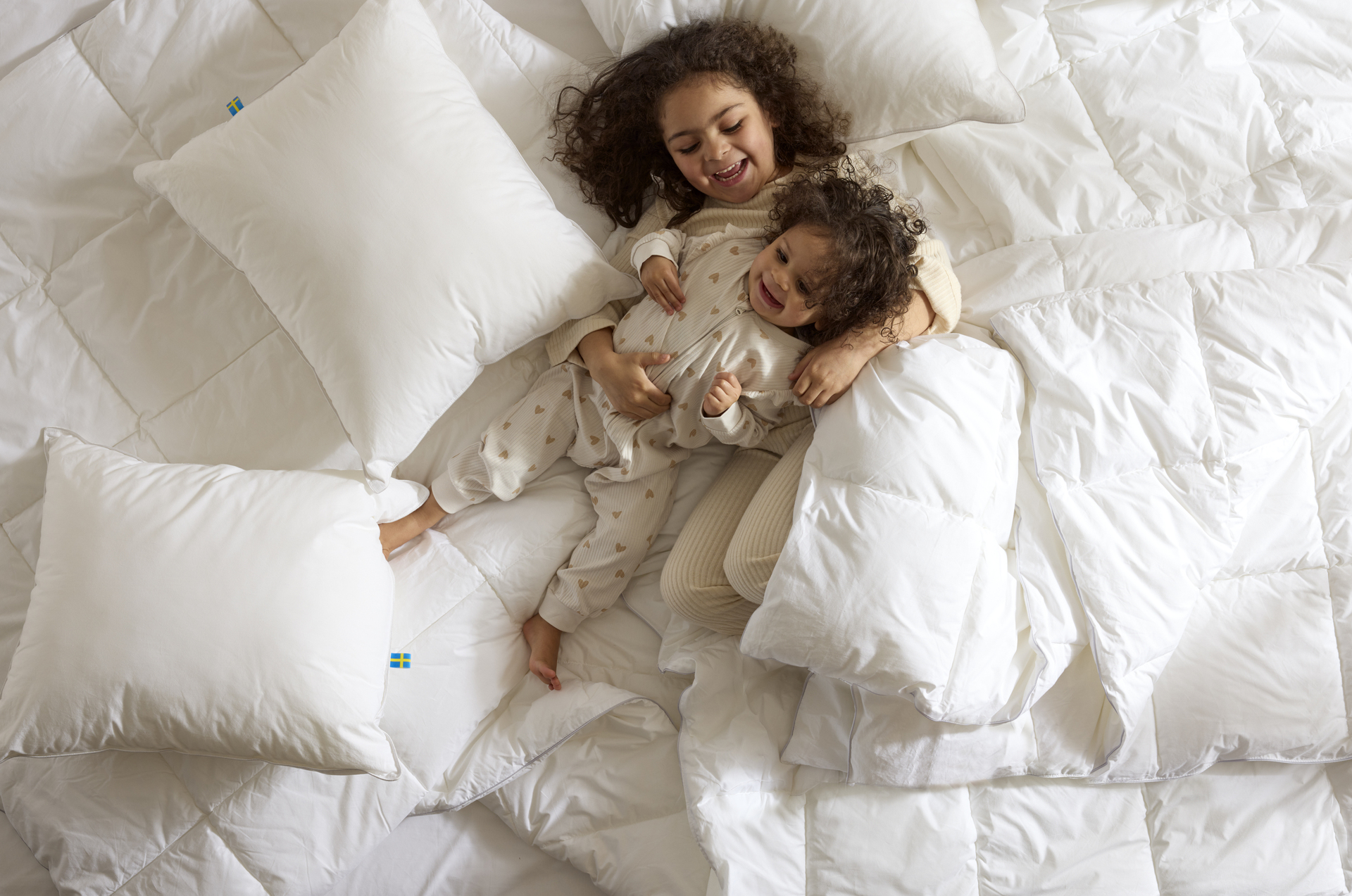 Två små barn busar bland täcken och kuddar i en obäddad säng.