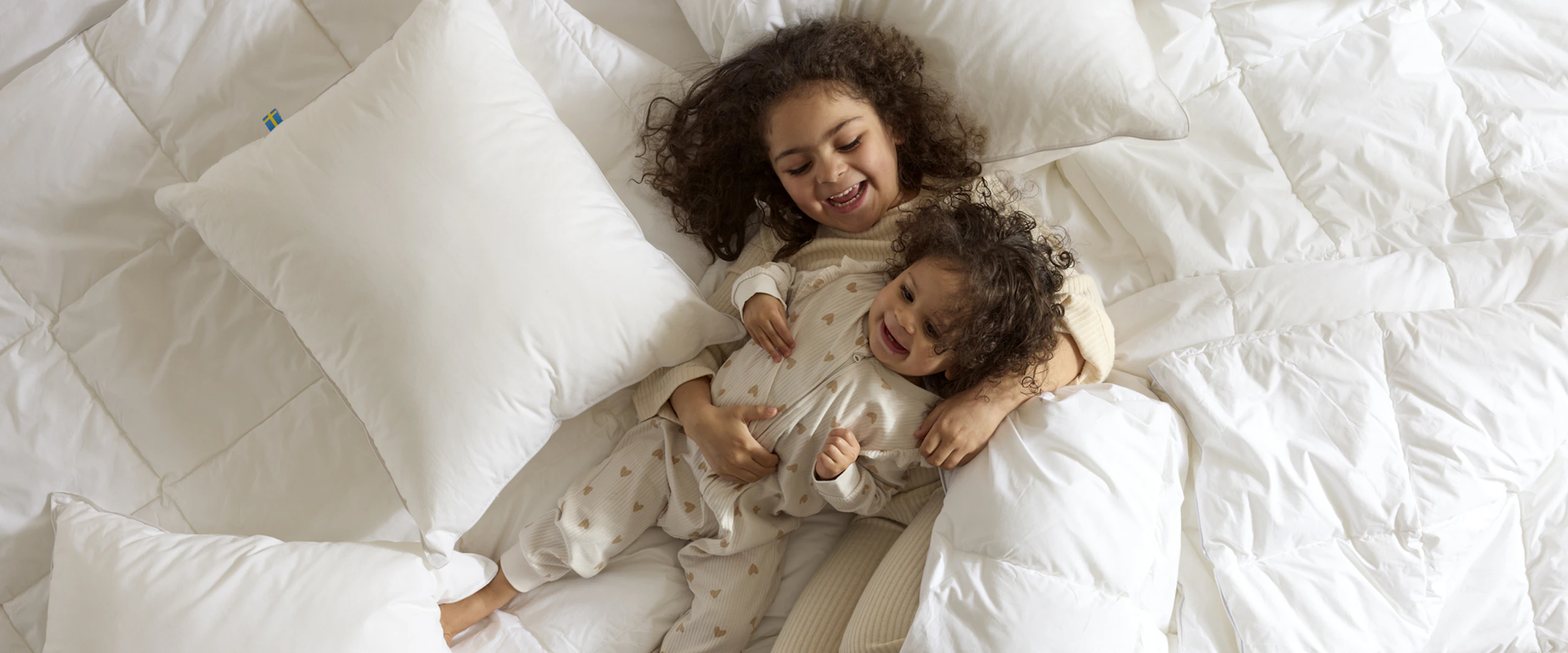 Två små barn busar bland täcken och kuddar i en obäddad säng.