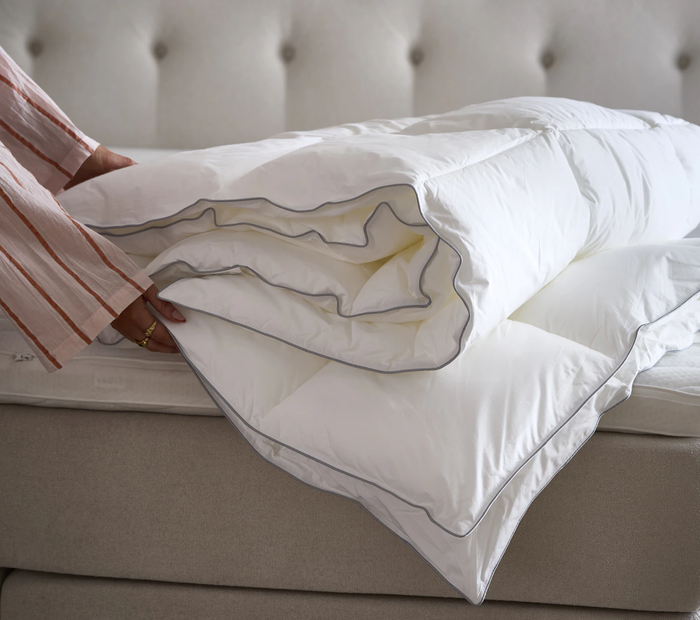 Två händer håller i ett fibertäcke utan påslakan som ligger på en säng.