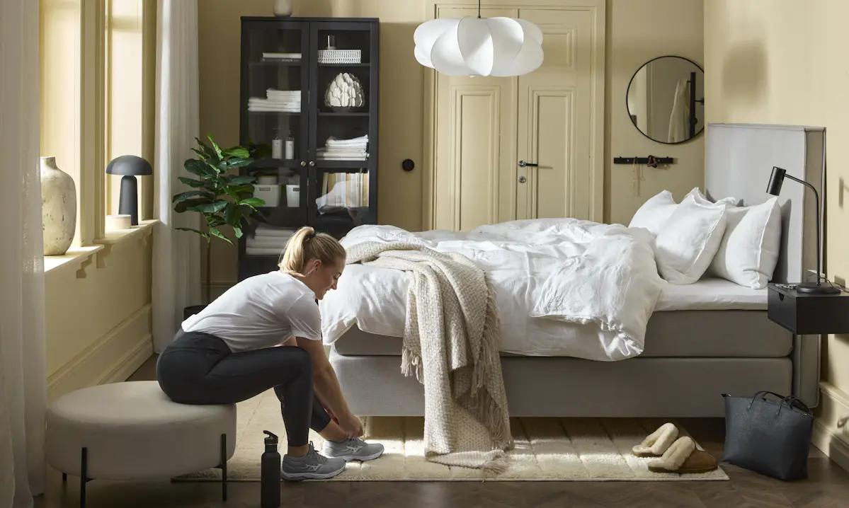 en tjej sitter på en pall och tar på sig skorna framför en grå kontinentalsäng i ett sovrum med gula väggar