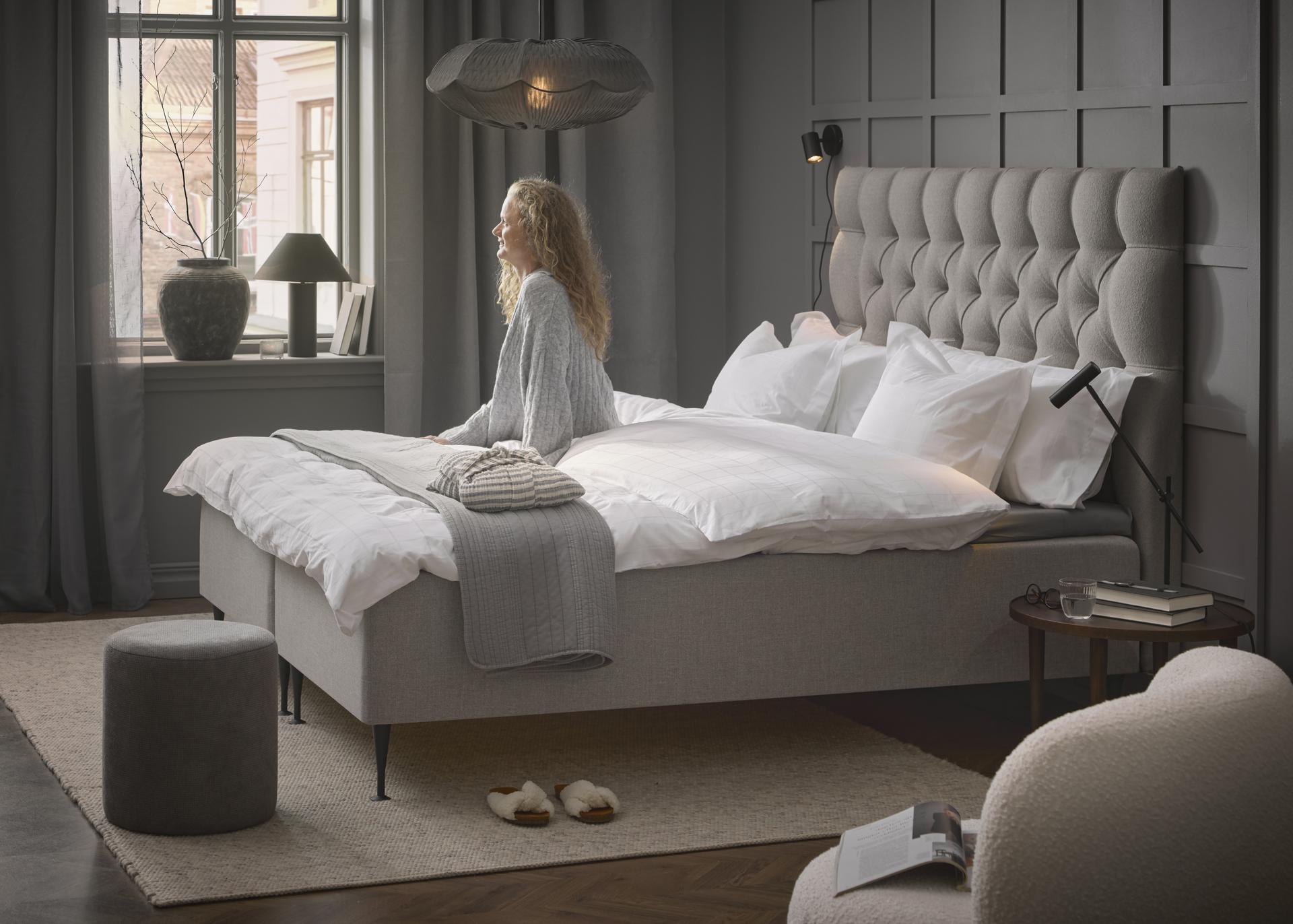 en tjej sitter i vita kläder på en beige kontinentalsäng och tittar ut genom fönstret