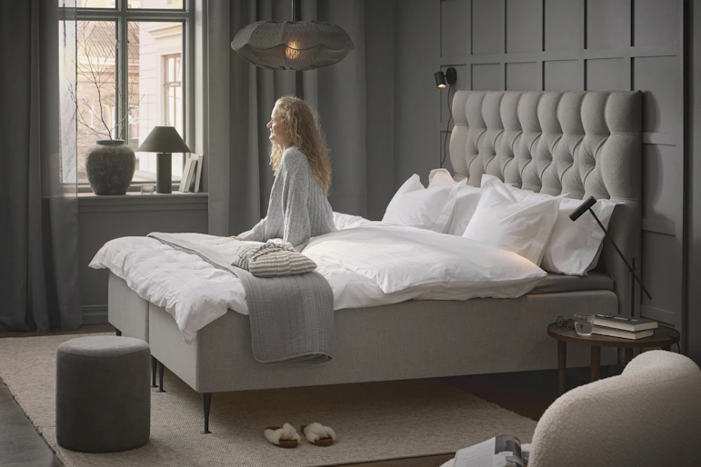 en tjej sitter i vita kläder på en beige kontinentalsäng och tittar ut genom fönstret