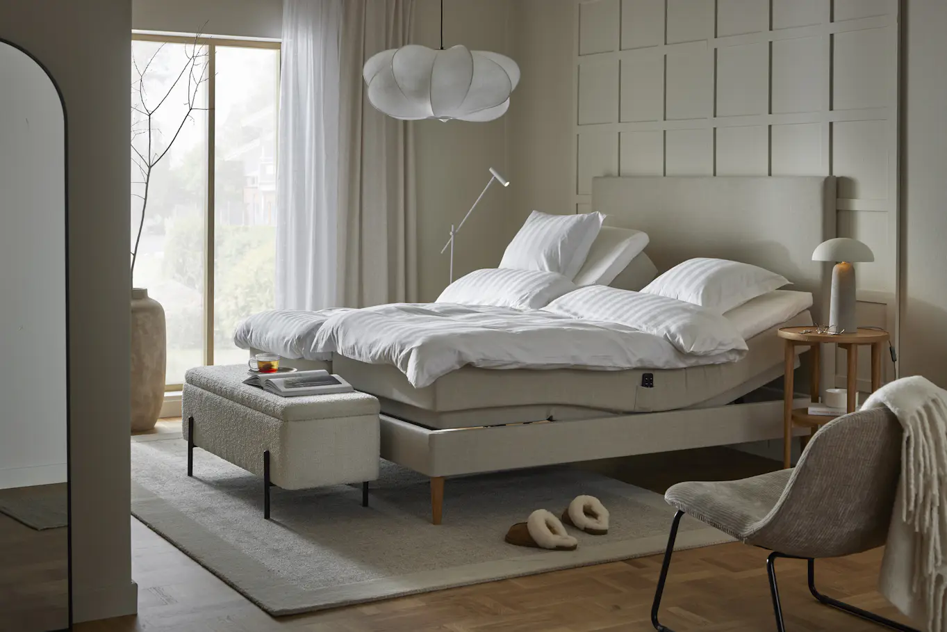 En beige ställbar säng står i ett beiget sovrum med stora fönster