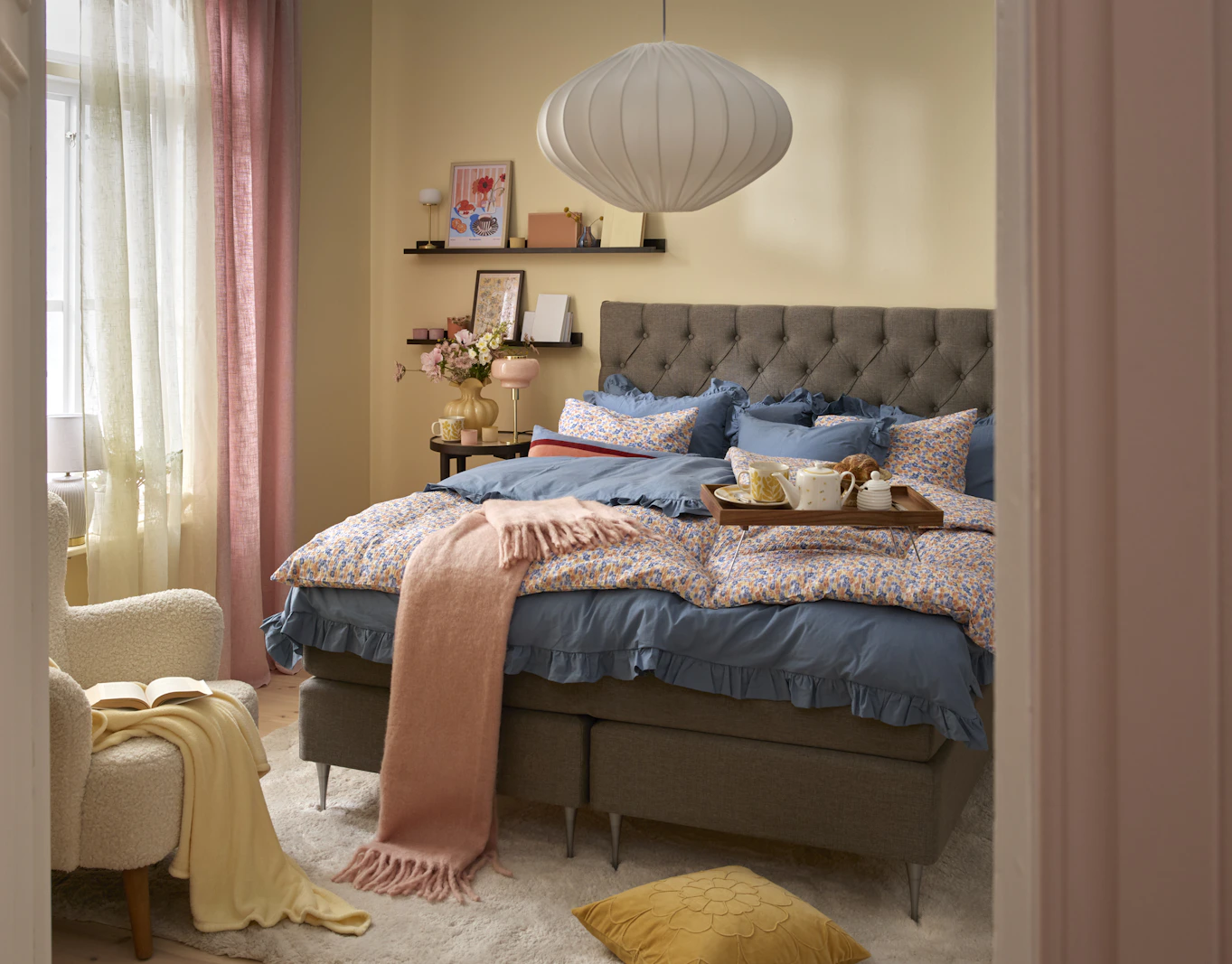 Kontinentalsäng i ett sovrum med påslakanset, plädar och kuddar i de vårliga färgerna gult, rosa, blått och nyanser av grått. 