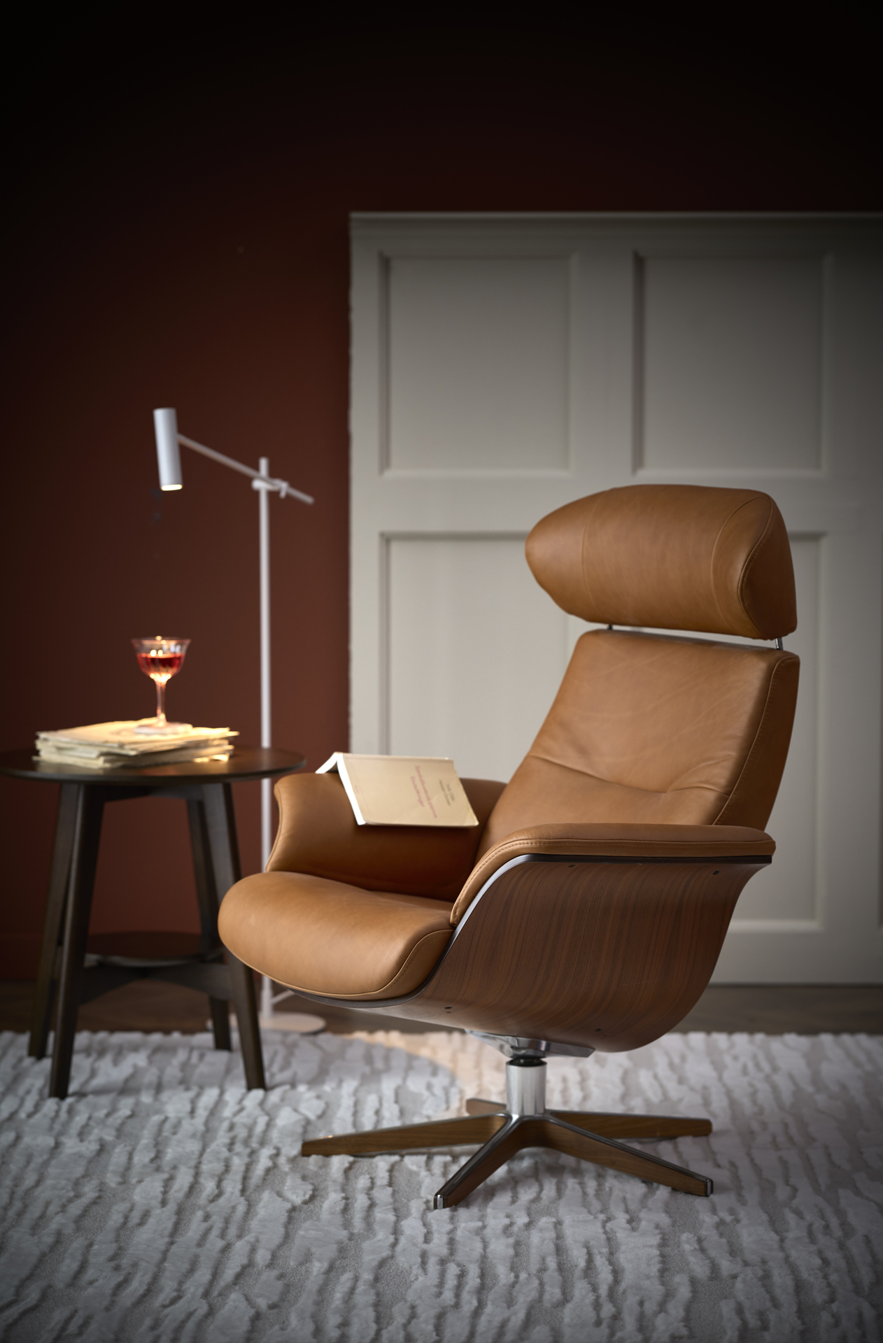 I ett rum med vit bröstpanel står en konjaksfärgad skinnfåtölj med underrede i valnöt brevid ett sidobord
