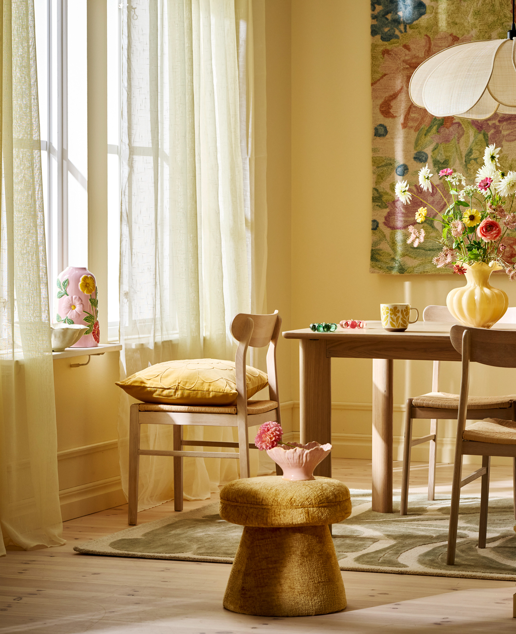 Ljus sipprar in genom tunna gardiner in i rum med gula väggfärger med matbord med trendig vas med blommor i. 
