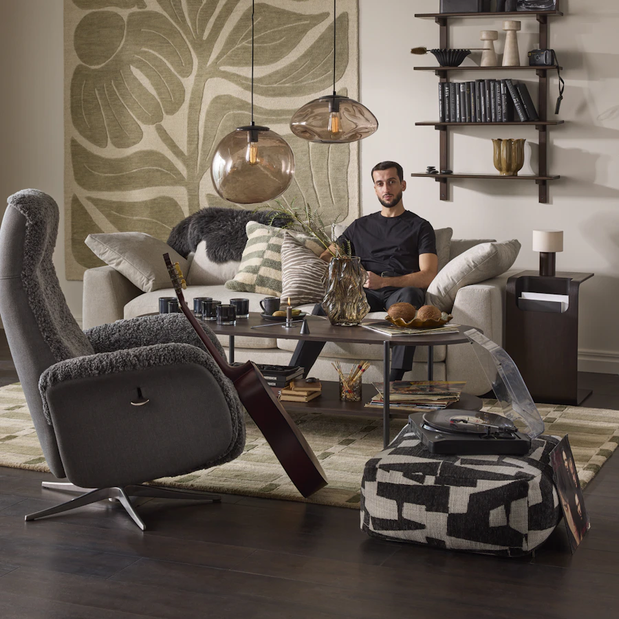 Vardagsrumsmiljö med fåtölj i förgrunden, i bakgrunden sitter en man i ljusgrå soffa.