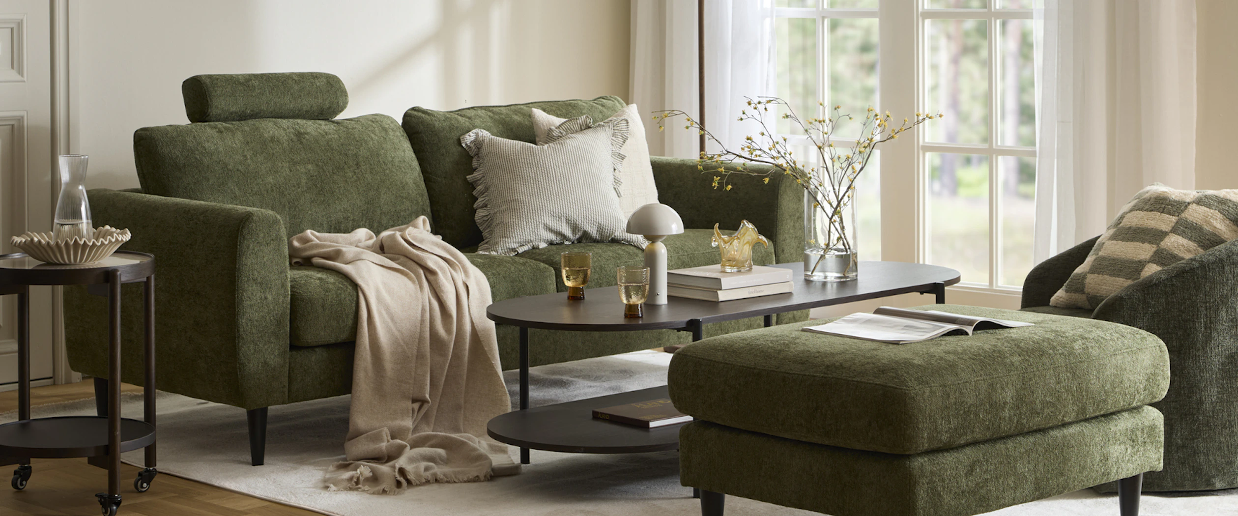 I ett ljust vardagsrum står en grön soffa vid ett soffbord i mörkt trä. Intill står en grön fotpall och en grön fåtölj