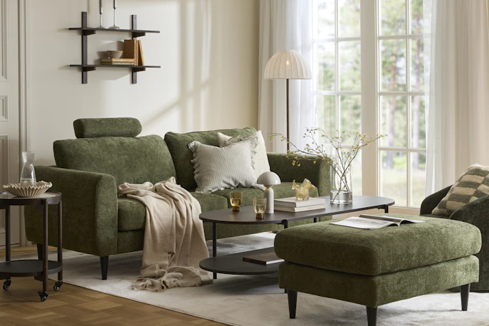 I ett ljust vardagsrum står en grön soffa vid ett soffbord i mörkt trä. Intill står en grön fotpall och en grön fåtölj