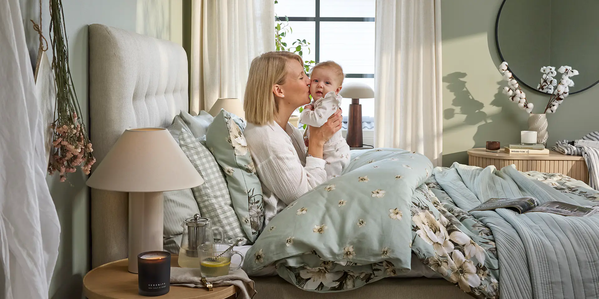 En blond tjej sitter nedbäddad i en säng och håller upp en bebis