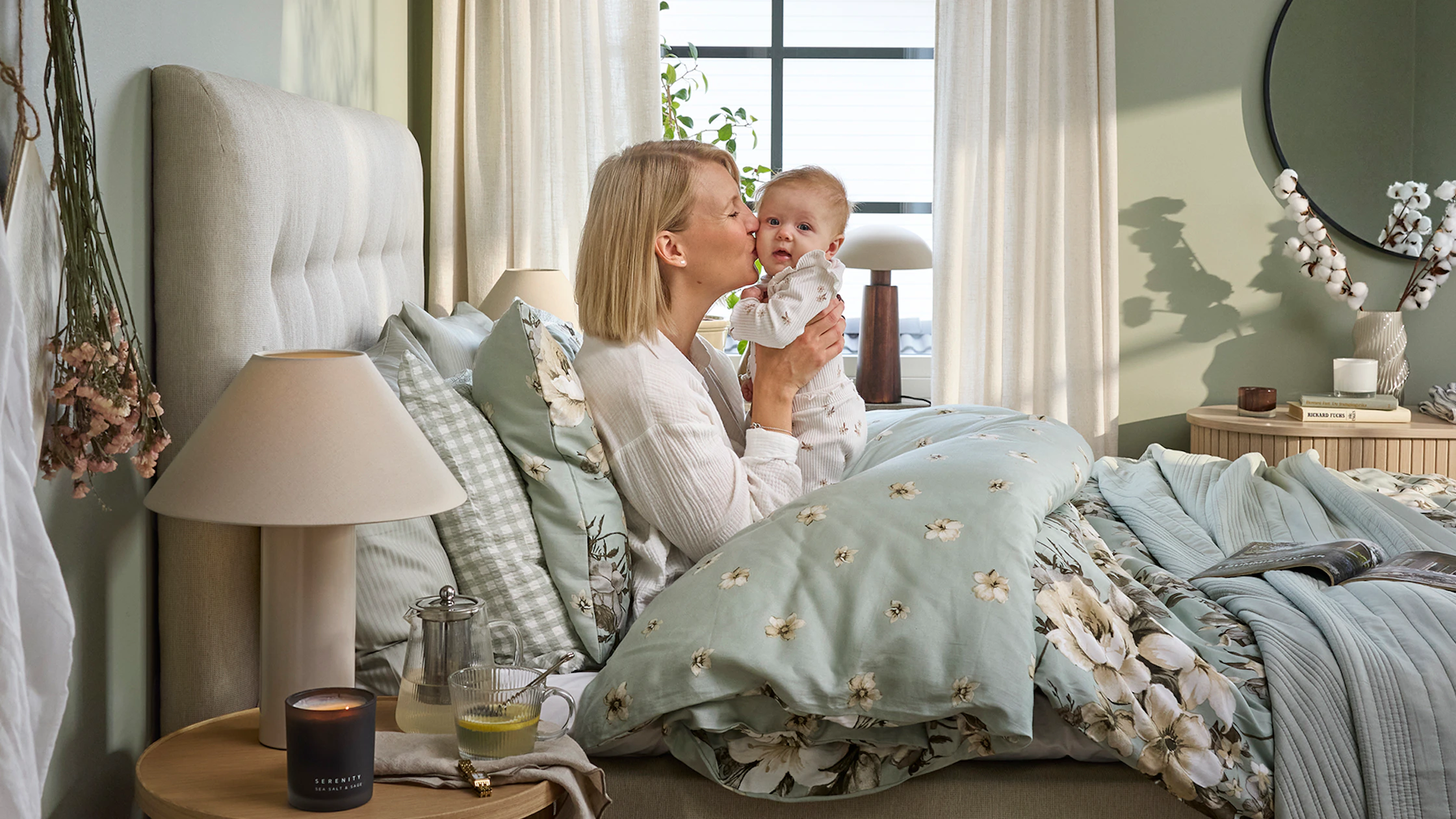 En blond tjej sitter nedbäddad i en säng och håller upp en bebis