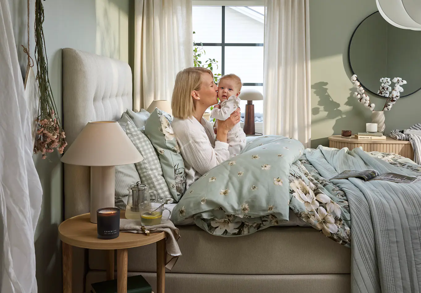 En blond tjej sitter i en säng och håller upp en liten bebis