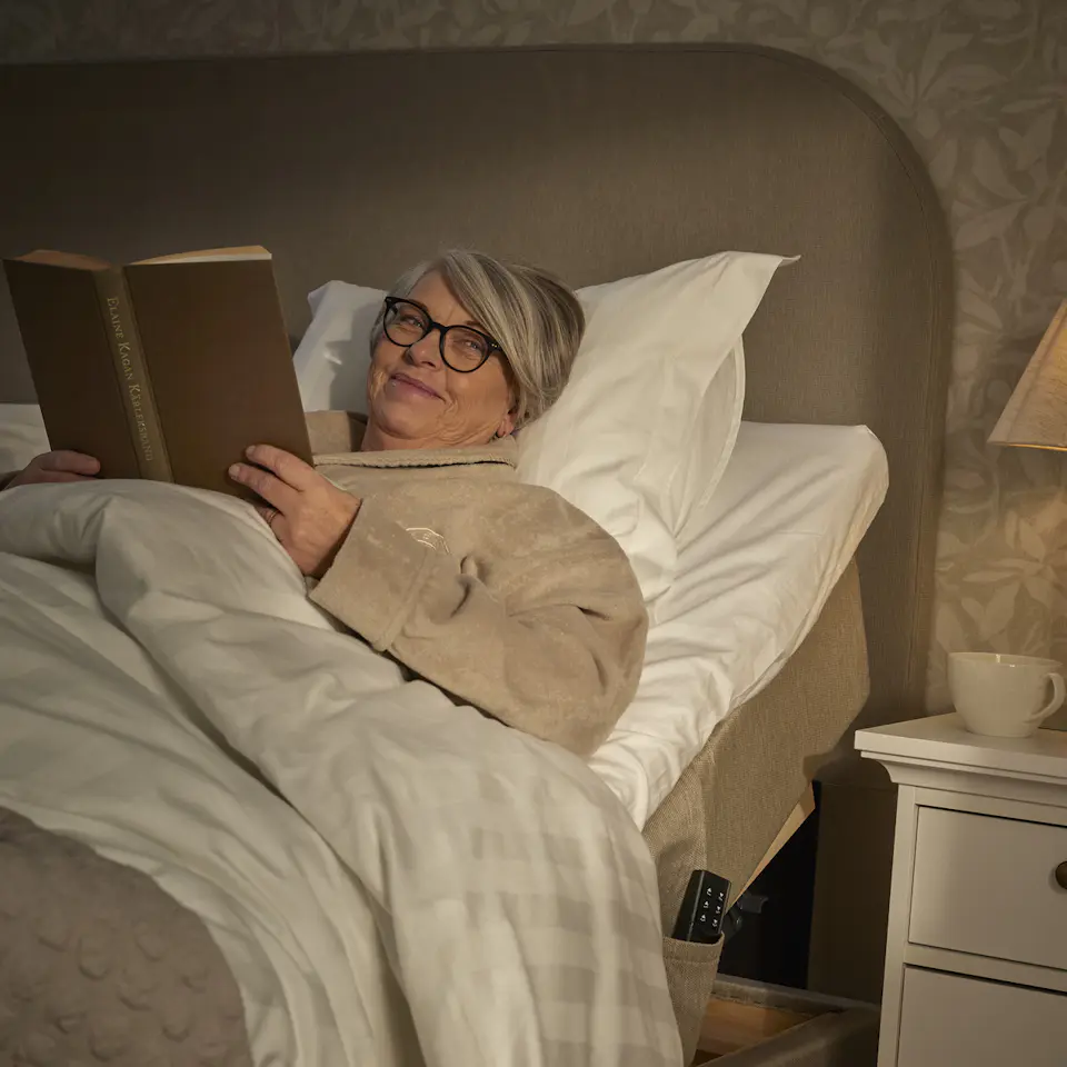 en kvinna ligger nedbäddad i en beige ställbar säng och läser en bok