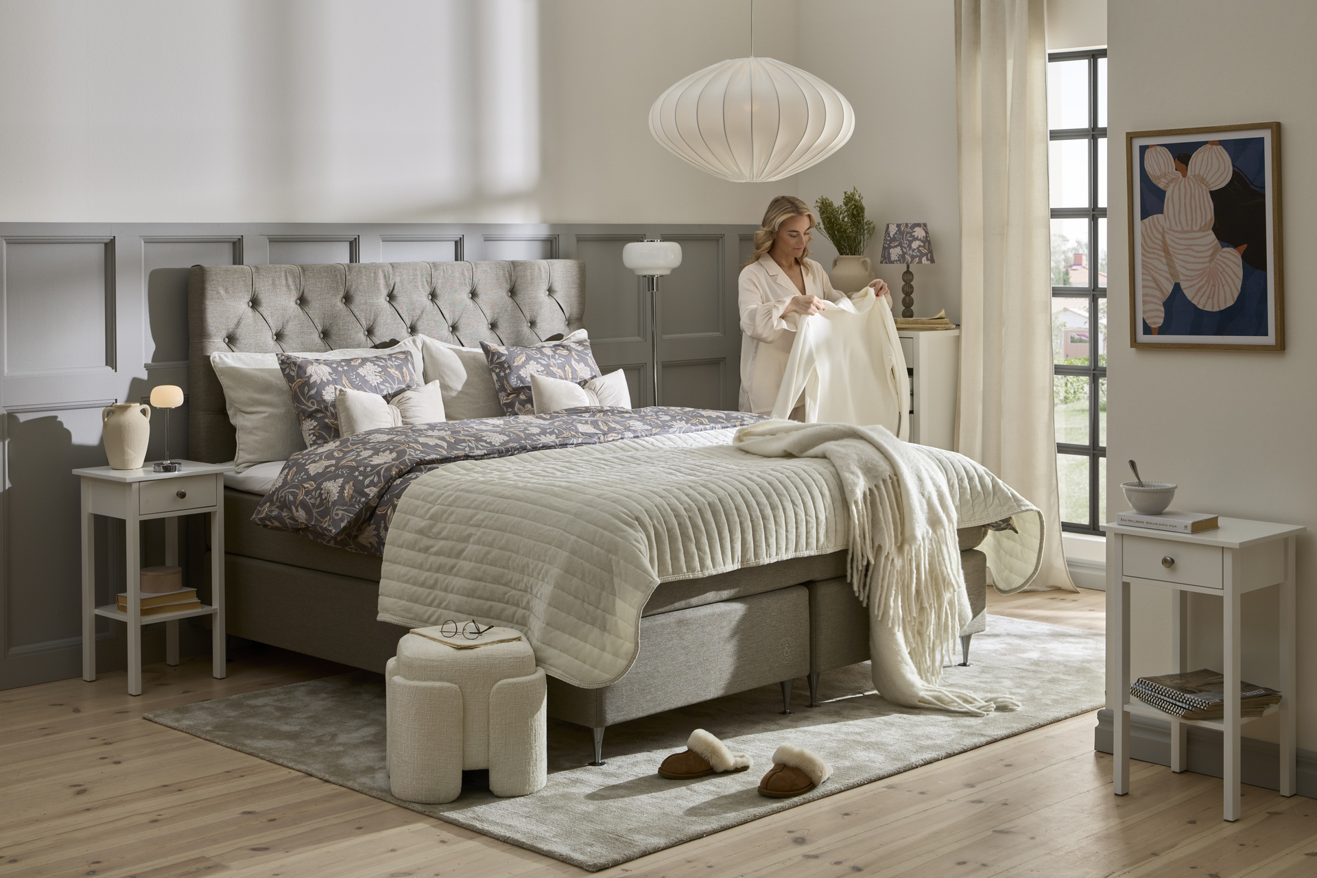 en blond tjej står i ett sovrum bredvid en grå kontinentalsäng och håller i en vit skjorta