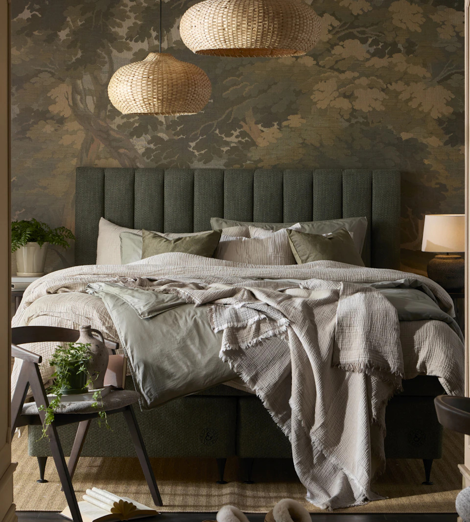 en säng med grön sänggavel och grå och bruna sängkläder står i ett sovrum med en mönstrad grön tapet