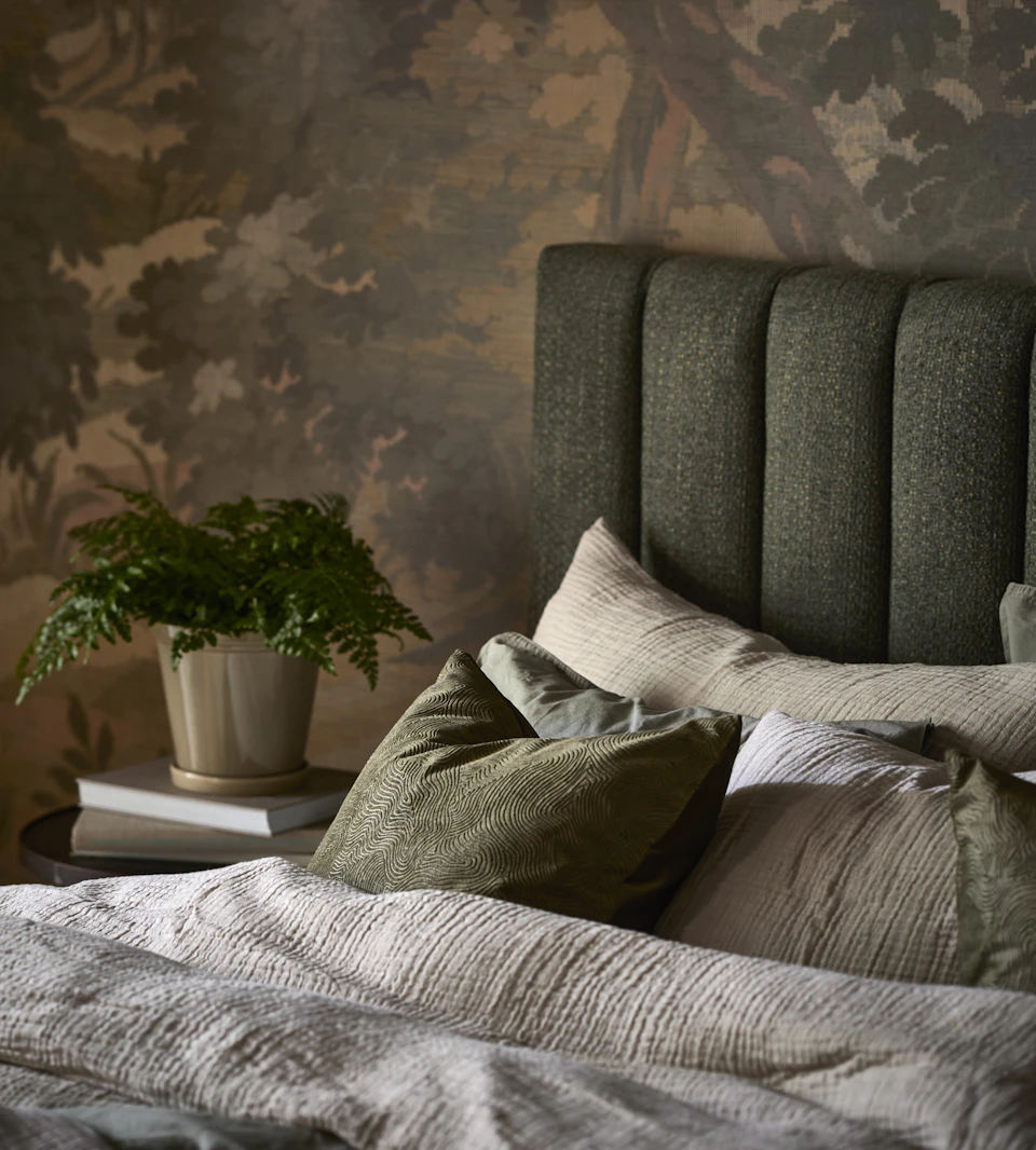 en säng med grön sänggavel och grå och bruna sängkläder står i ett sovrum med en mönstrad grön tapet