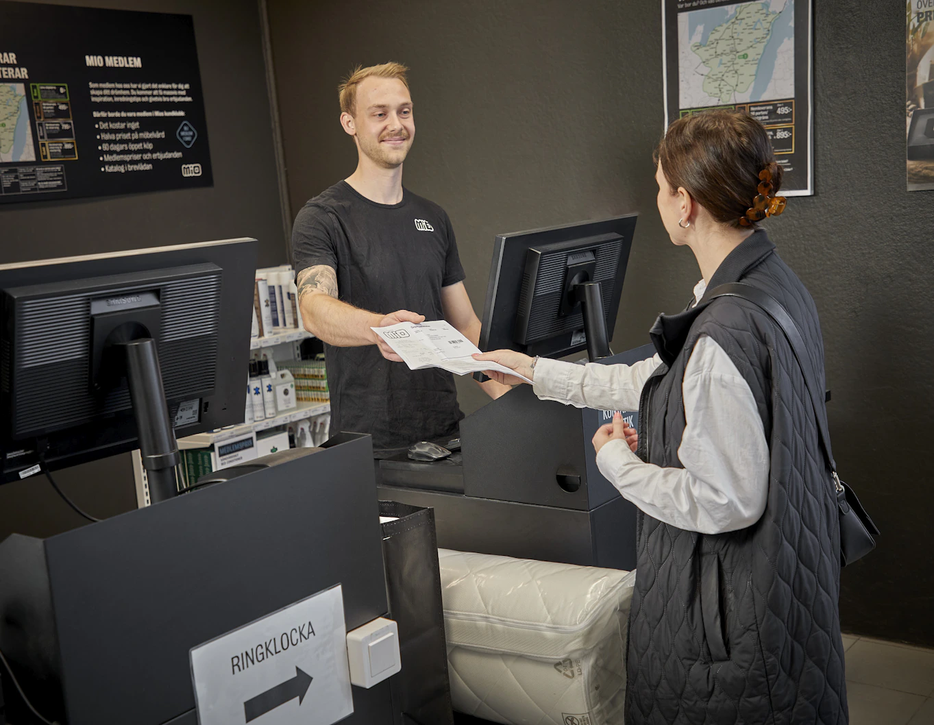 Butiksmedarbetare med svart t-shirt ler och överlämnar kvitto till kund vid varuuutlämningen på Mio. 