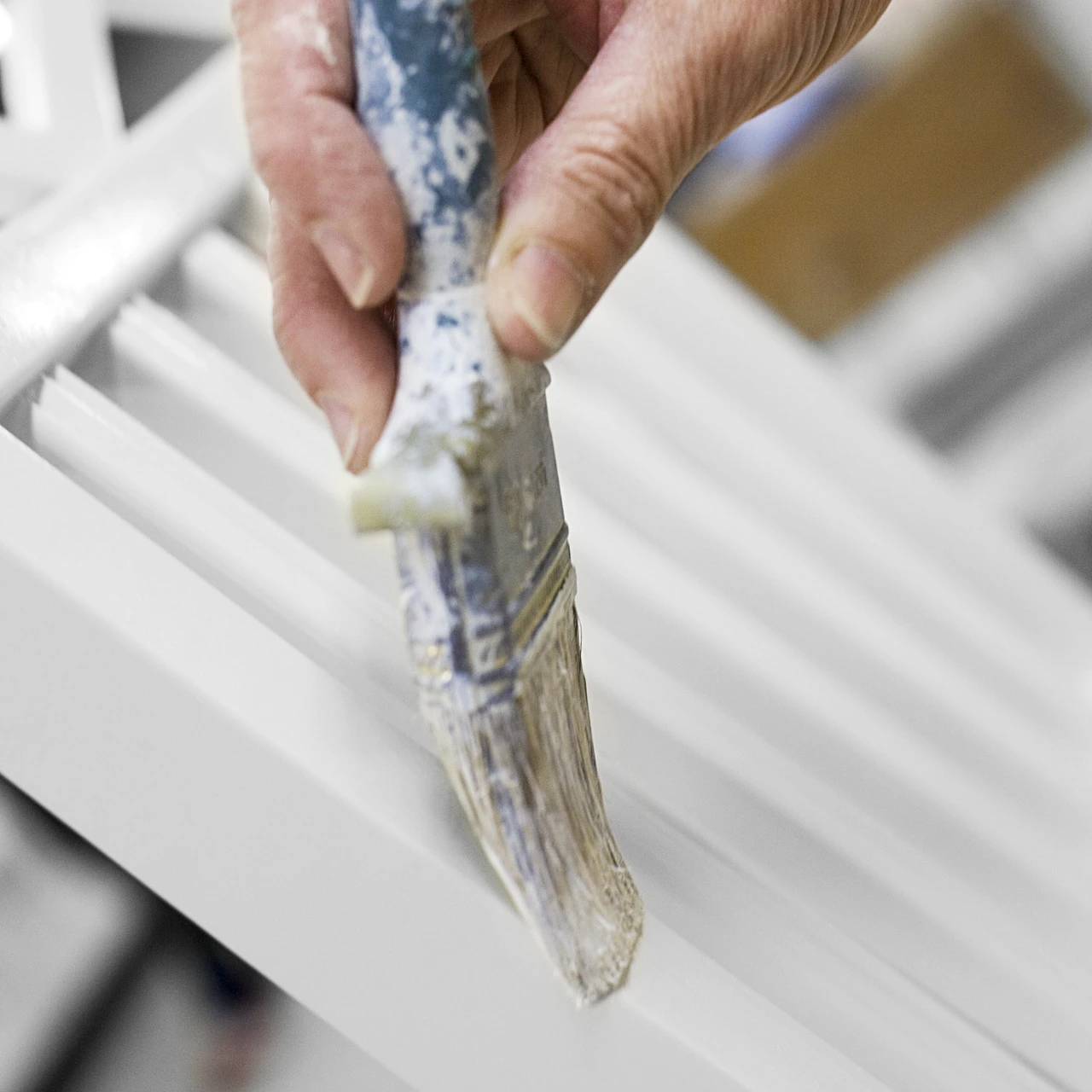 Närbild på hand som stryker pensel med vit färg över ribborna på en stolsrygg
