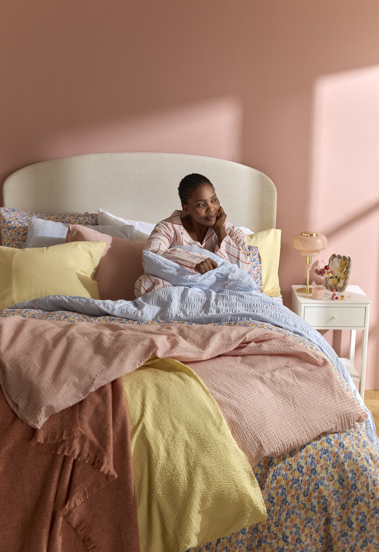 Kvinna sitter upp i säng med färgglada sängkläder i vårliga färger och ljusrosa vägg.