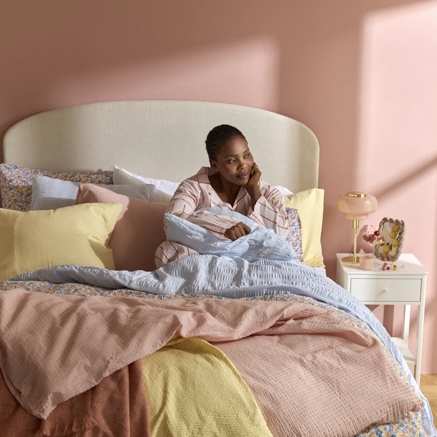 Kvinna sitter upp i säng med färgglada sängkläder i vårliga färger och ljusrosa vägg.